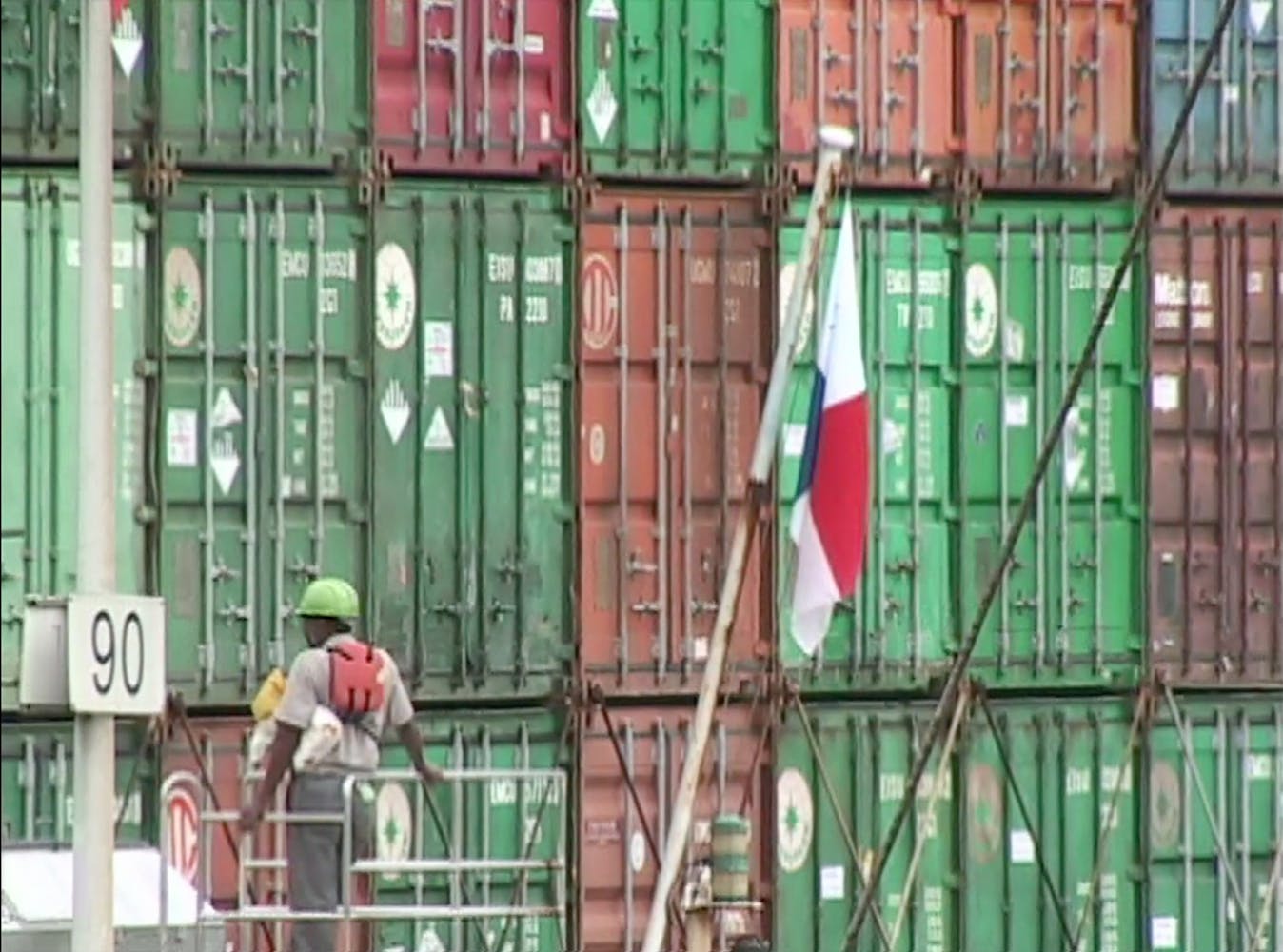 Still image of green, orange and red shipping containers stacked together, filling the frame. A worker stands looking on.