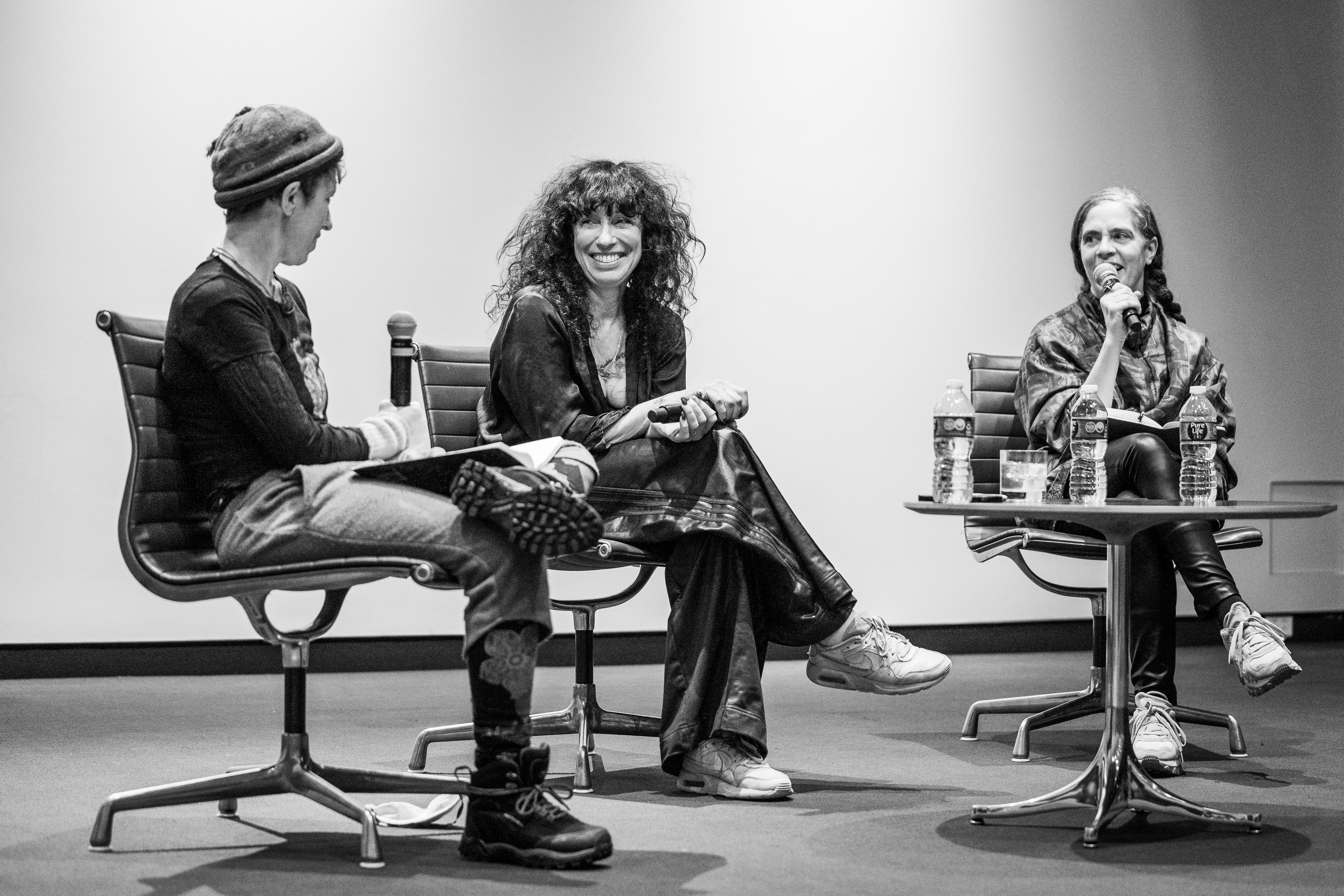 Three women sit on chairs on a stage holding microphones, talking, and laughing.