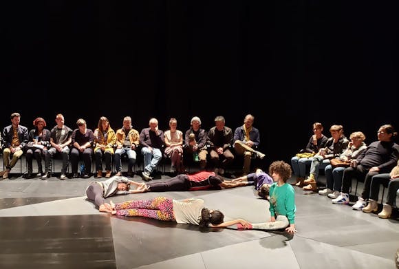Five dancers lying on the stage floor in a triangular pattern with audience members seated around them