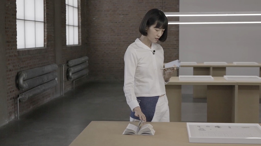 A woman dressed in white reads from a small book above a table with framed drawings and another book.