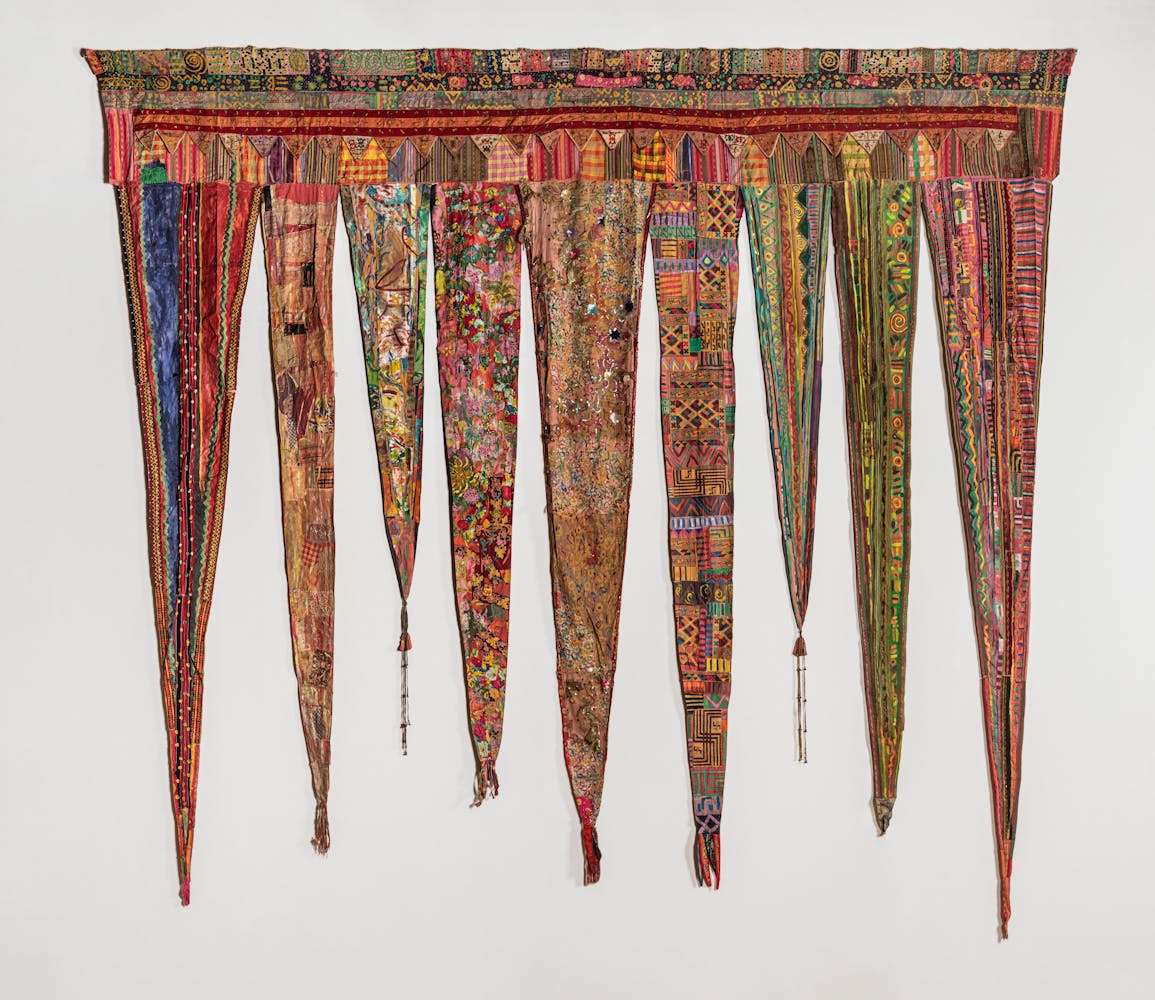 A colorful artwork of long trinangluar fabric pieces made up of many different fabrics and patterns hangs on a wall.