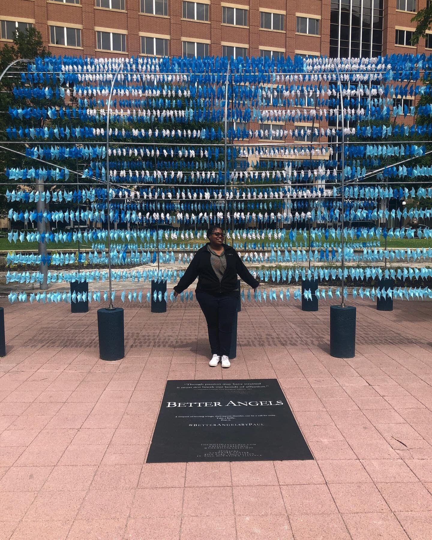A dark skinned woman is standing in front of an installation. the installation has horizontal bars with hundreds of pinwheels on them. They are mostly blue with some white. There is a sign on the ground with the words "better angels" visible