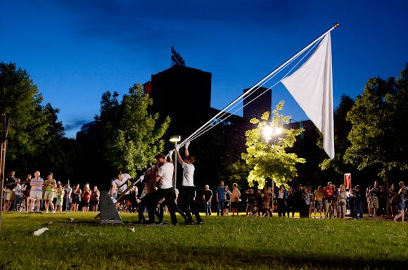 Five people in white shirts and dark pants raising a large white flag, in a field, surrounded by an audience, at night