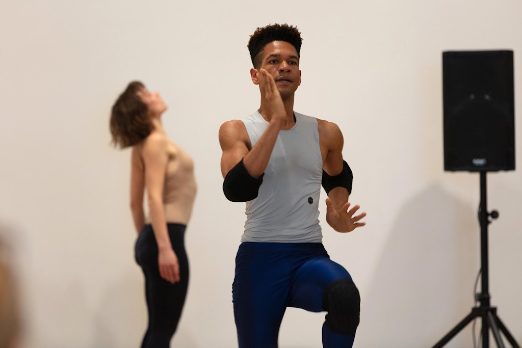 Two dancers in a white gallery. In the foreground, a male dancer in a gray tank top and elbow pads is running in place. In the background, a female dancer stands with her upper back arched. On the right side of the frame, there is a single speaker on a stand.