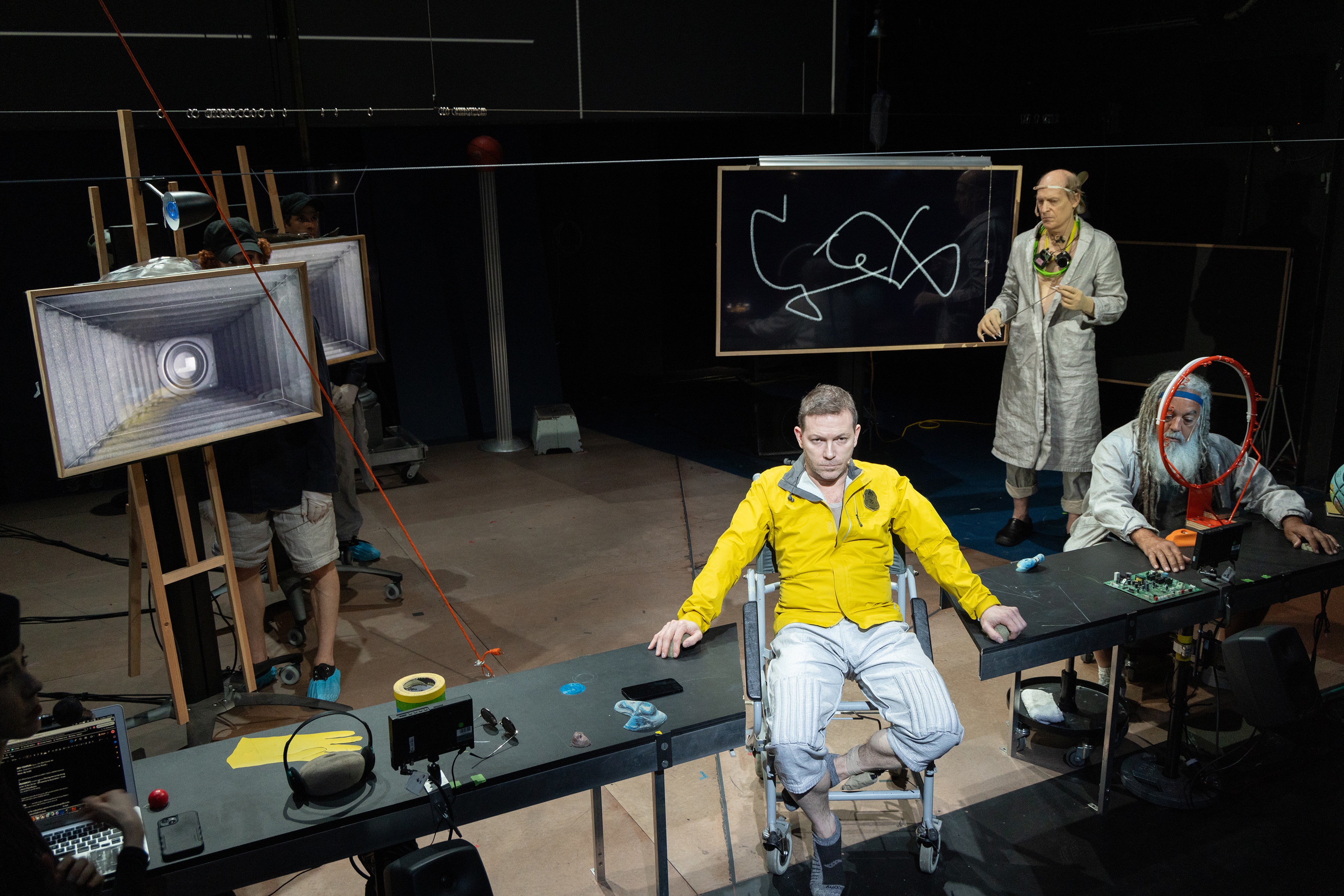 Five performers with light skin pose in a dark, cluttered set with a blackboard, table, and speakers.