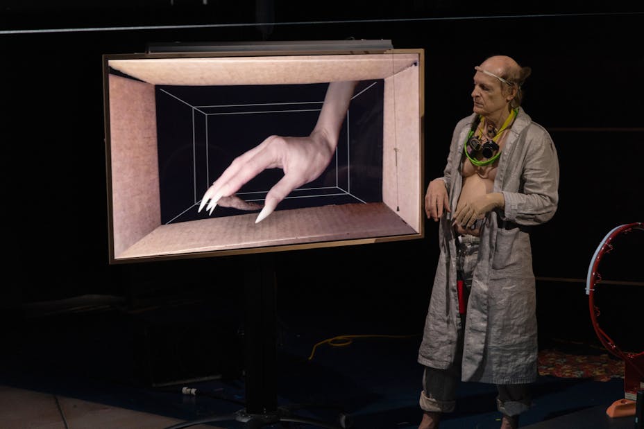 An older light-skinned man looks at a giant hand with long fingernails in a box.
