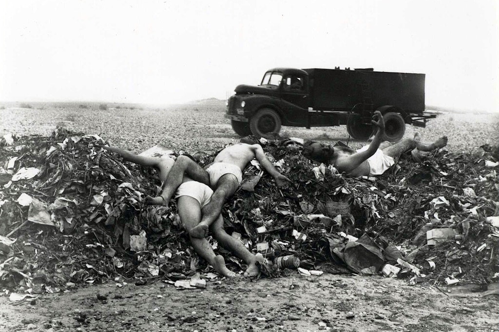 3 men, apparently dead, laying on top of a pile of garbage wearing only underwear. Truck in background.