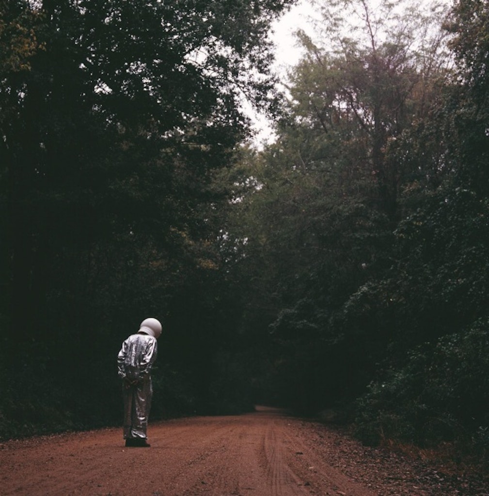 Man in space suit on a dirt road in woods.