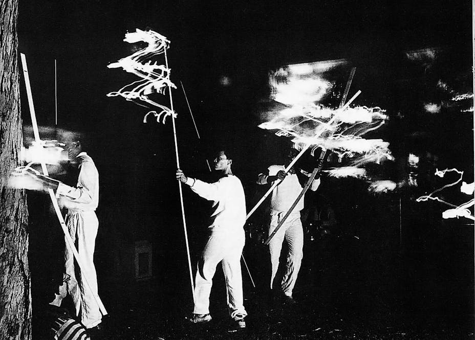1982 black and white image of artists wearing white and carrying long sticks, performing "Flying"