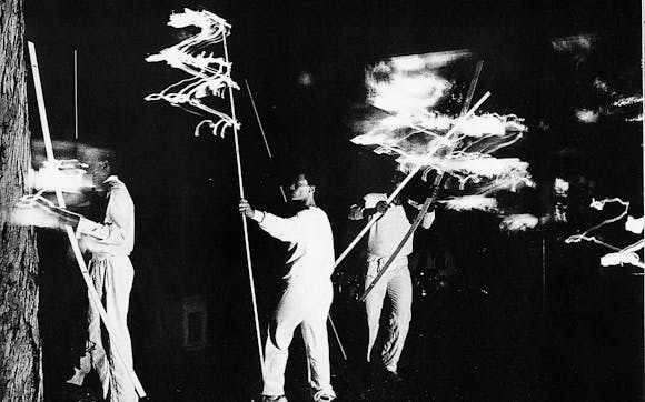1982 black and white image of artists wearing white and carrying long sticks, performing "Flying"