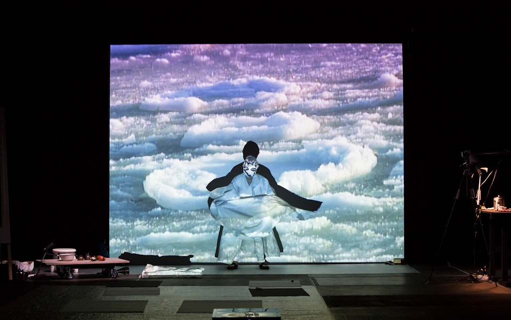 A person in a costume and mask stands on a stage in front of an images of ice or clouds.