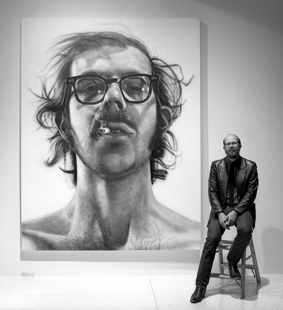 Man sitting on stool in front of large portrait of man wearing glasses with cigarette in mouth