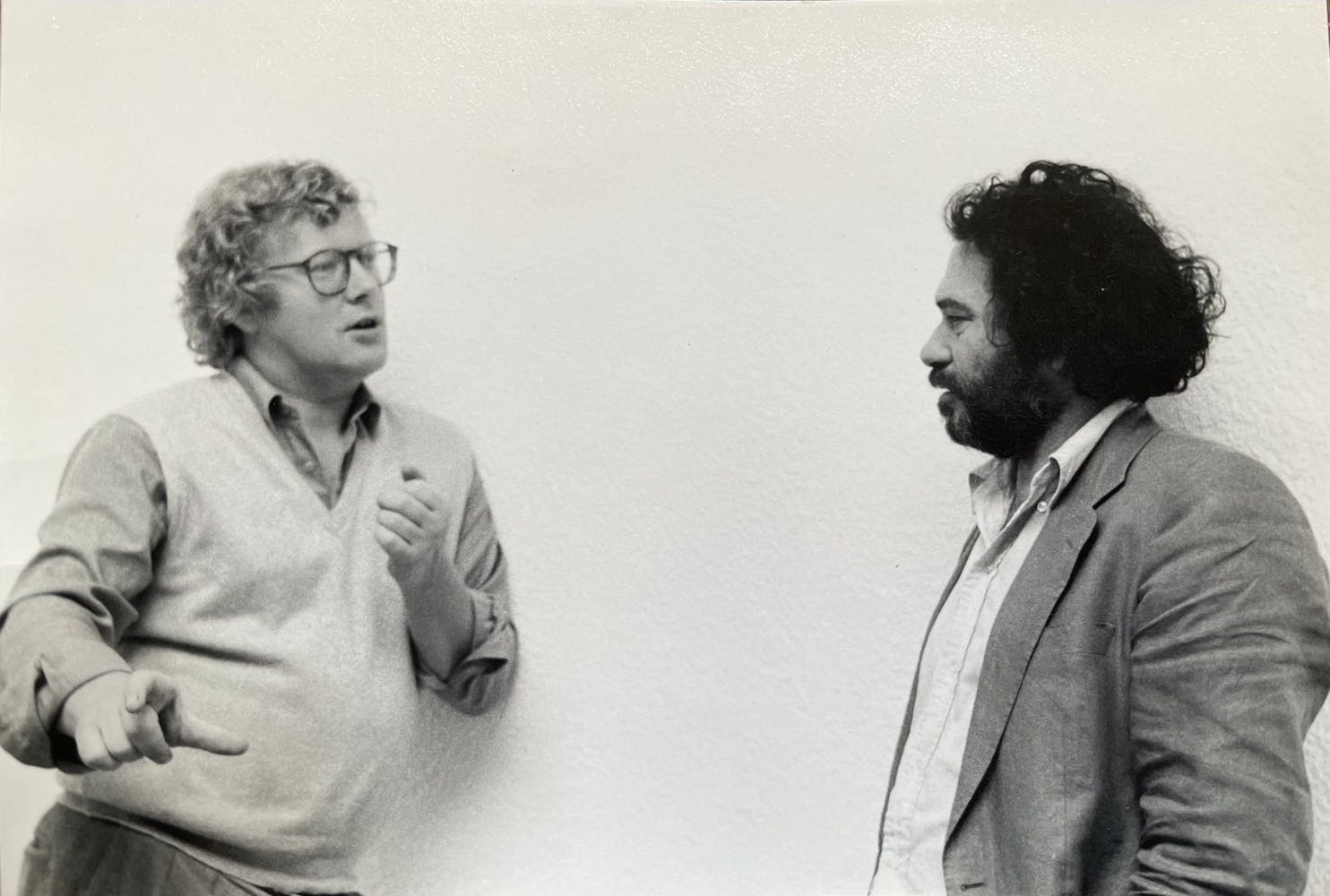 Two men stand talking next to a blank wall.