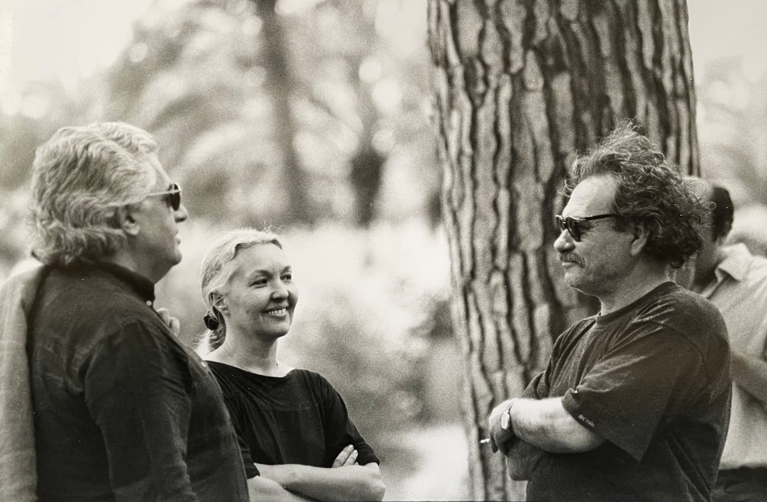Two men and a woman stand outside near trees talking.