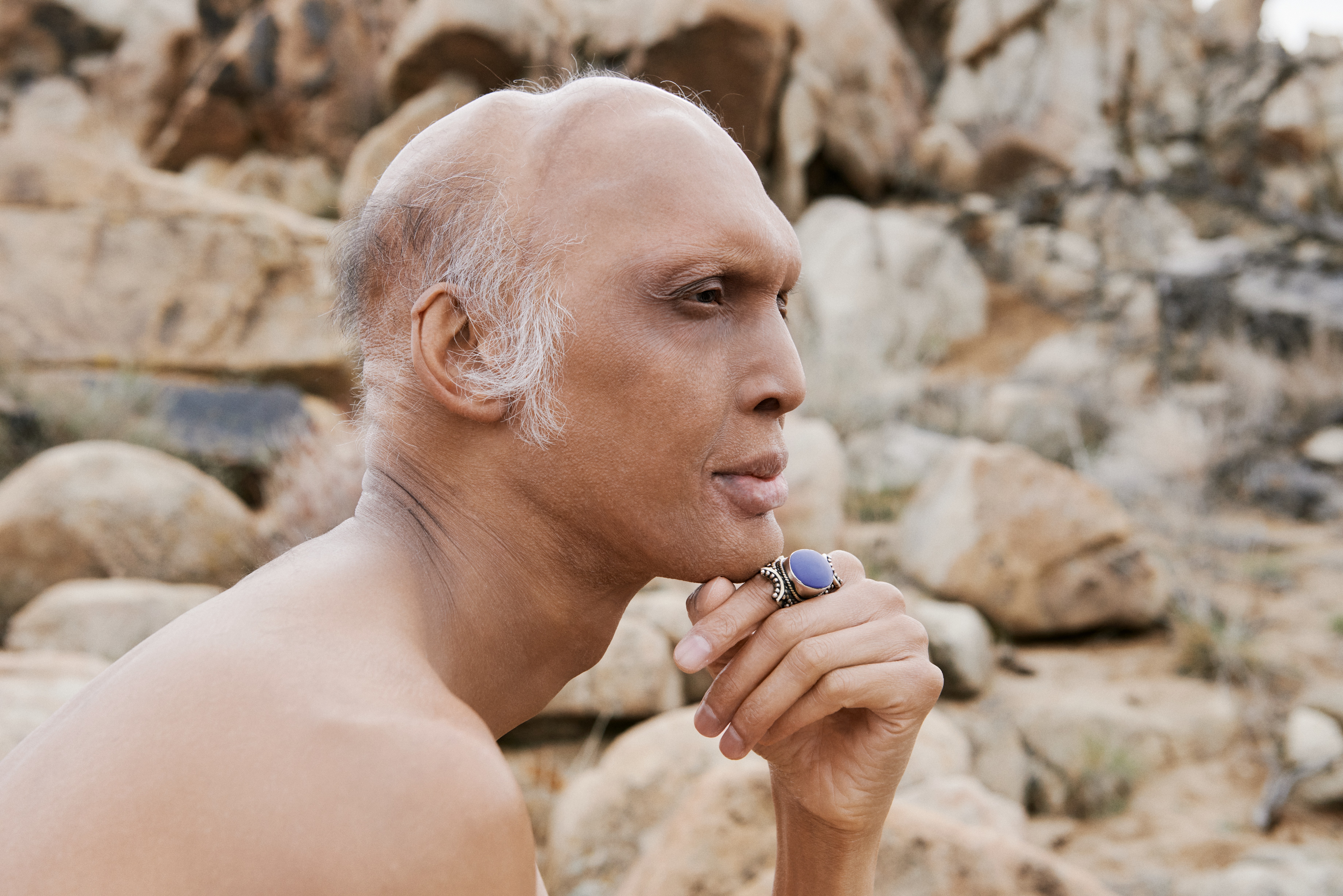 A person with medium skin rests their chin on their hand in front of a tan, rocky landscape.