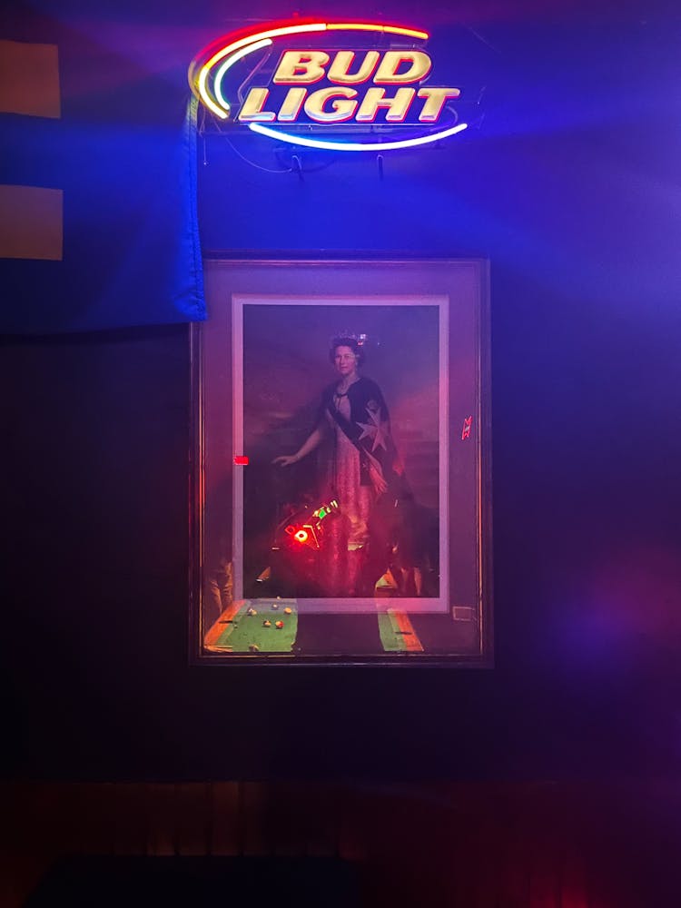 1. Photograph of a framed portrait of Queer Elizabeth under a neon Bud Light sign. A pool table can be seen in the reflection of the portrait’s glass.