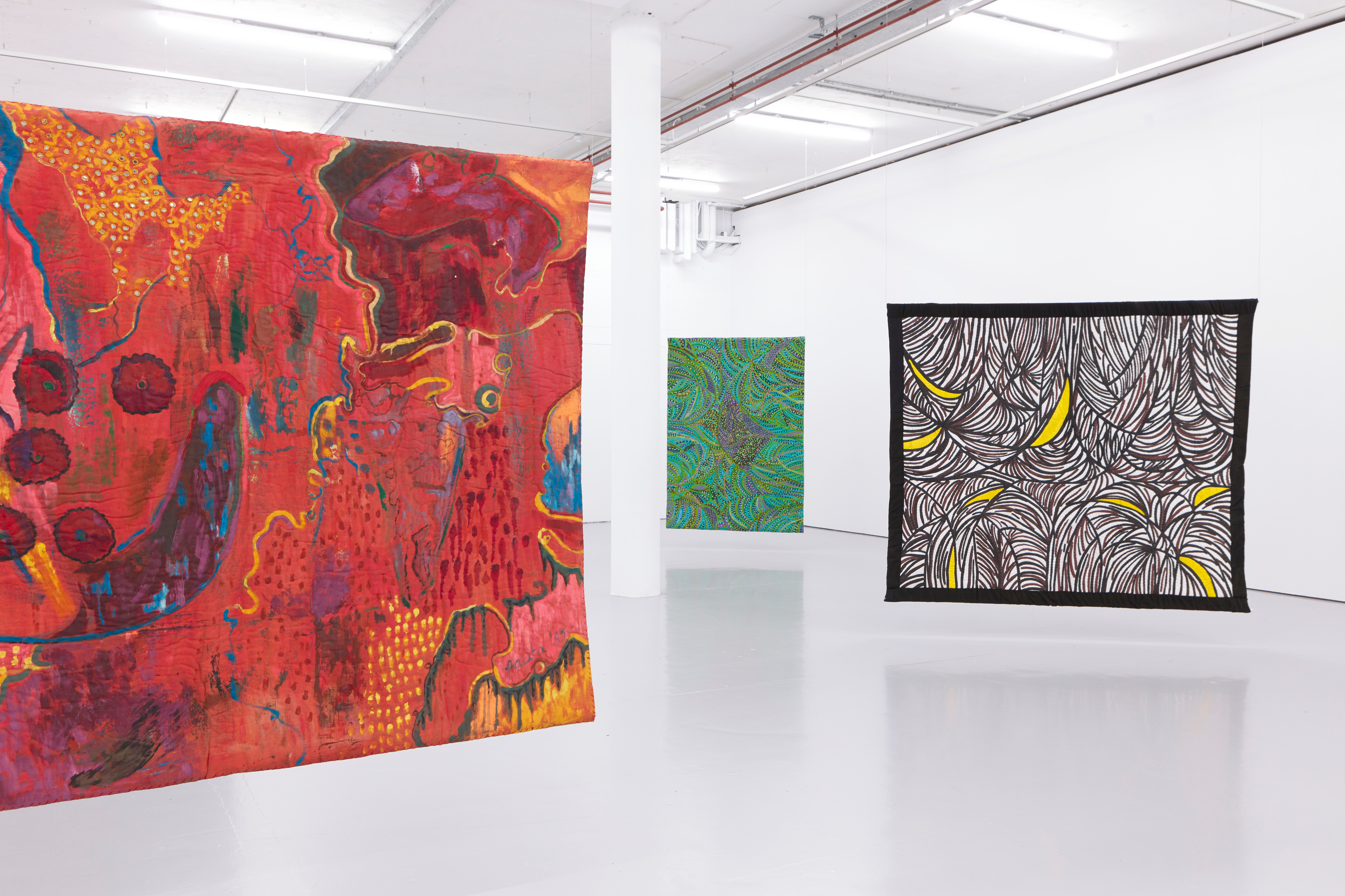 Large, colorful textile works hang in a large white art gallery.