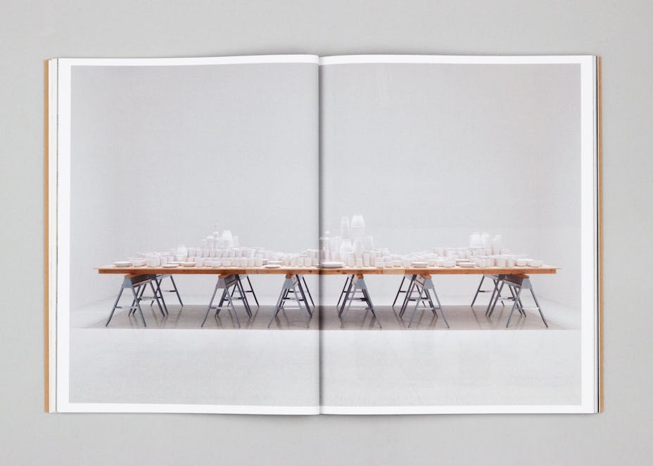 Two pages of a book with a large photograph of a table covered in a cityscape made of ceramic plates, cups, and bowls.