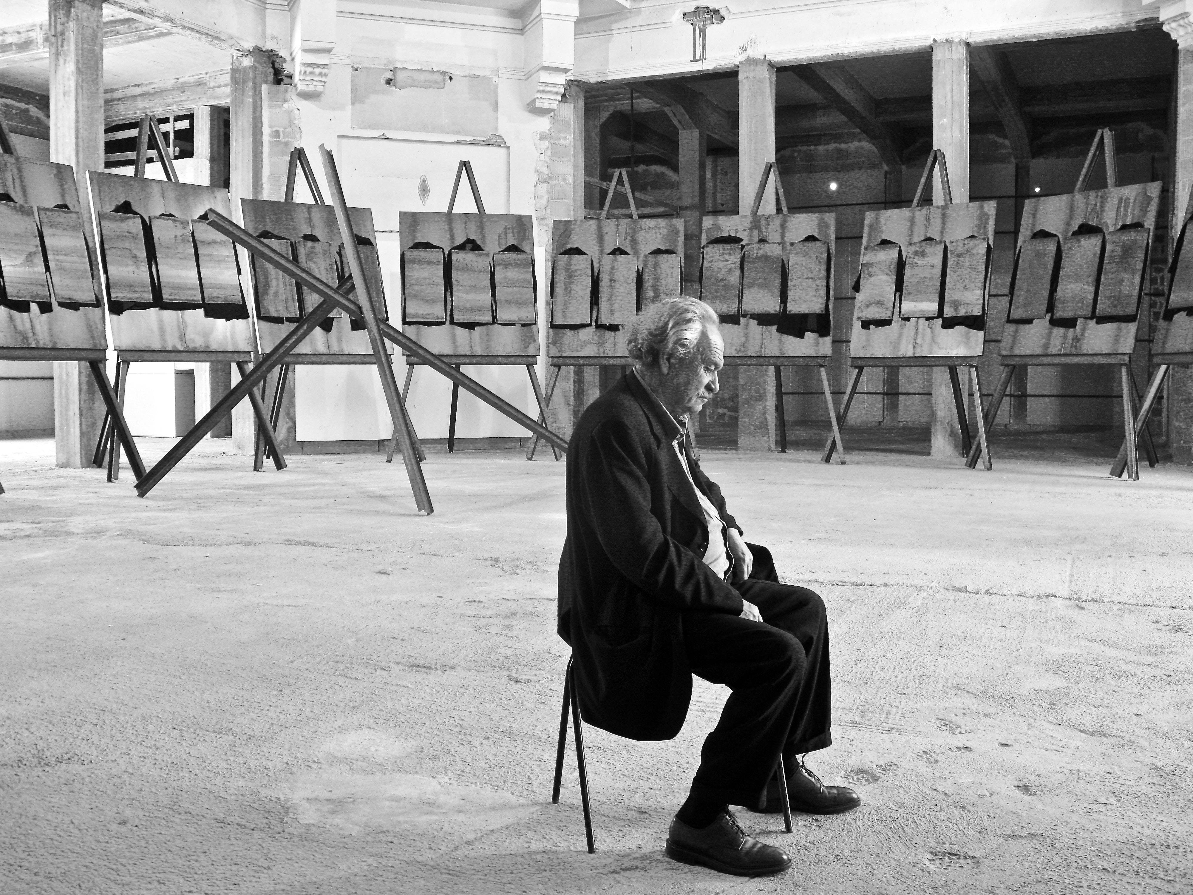 A man in a suit sits in a gallery surrounded by large paintings.