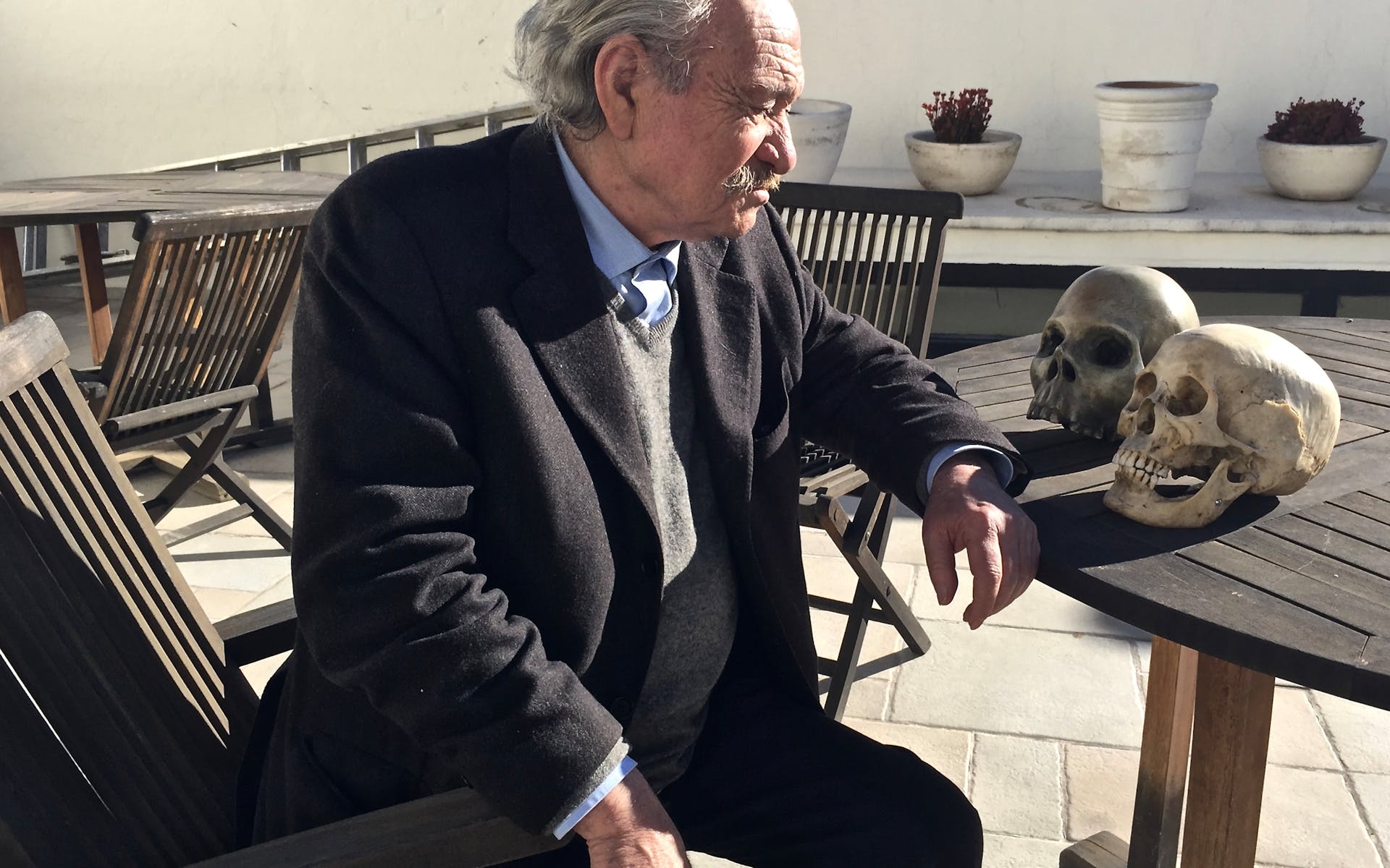 An older man in a suit sits gazing at two skulls on a table inf ront of him.