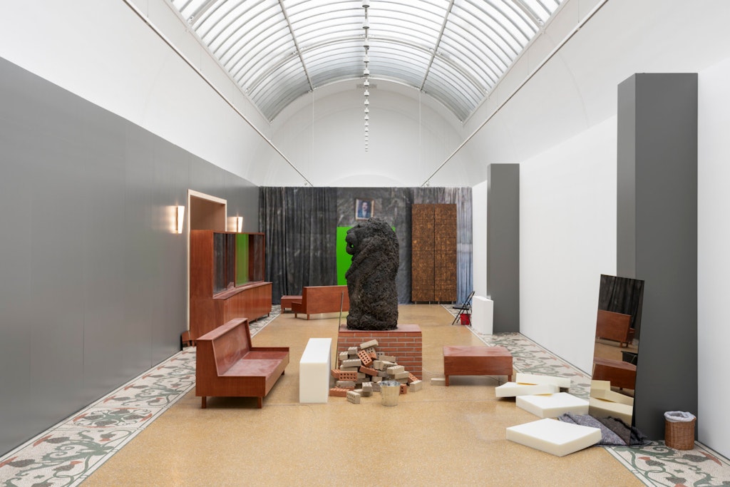 a long room with a barrel vaulted ceiling with many objects seemingly haphazardly strewn around the room. The edges of the room's floor are patterned in an abstracted vegetal design, a bench, a pile of bricks, a mirror, awaste basket, and a sculpture of a lion made from earth are all visible.