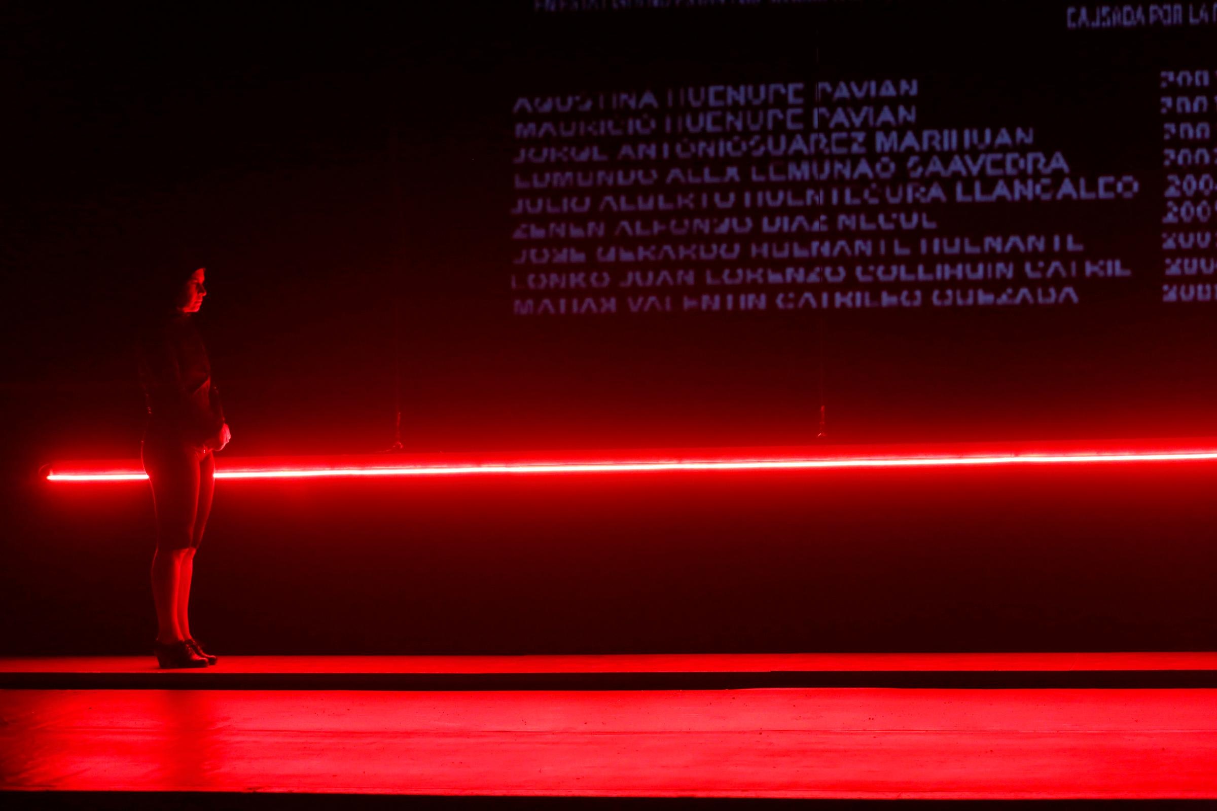A woman in black stands in front of a glowing red line with projections of names in white above it.