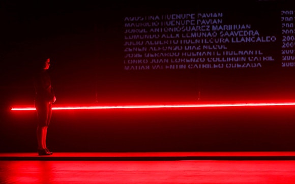 A woman in black stands in front of a glowing red line with projections of names in white above it.