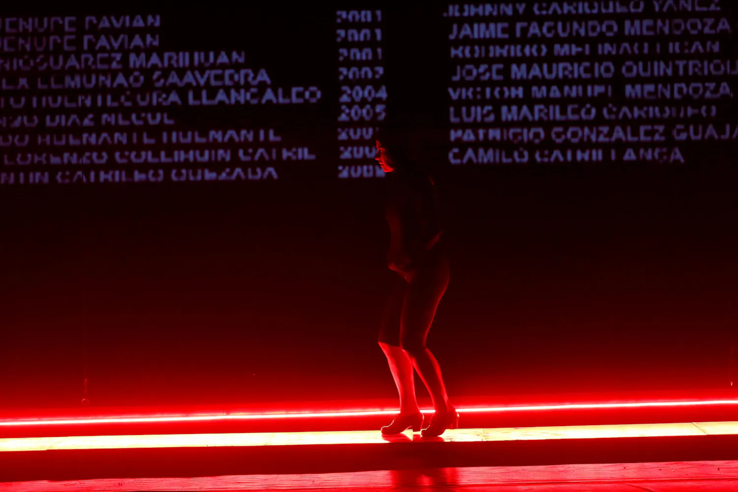 A woman dances on a black stage with red lights at her feet and a list of names projected behind her.