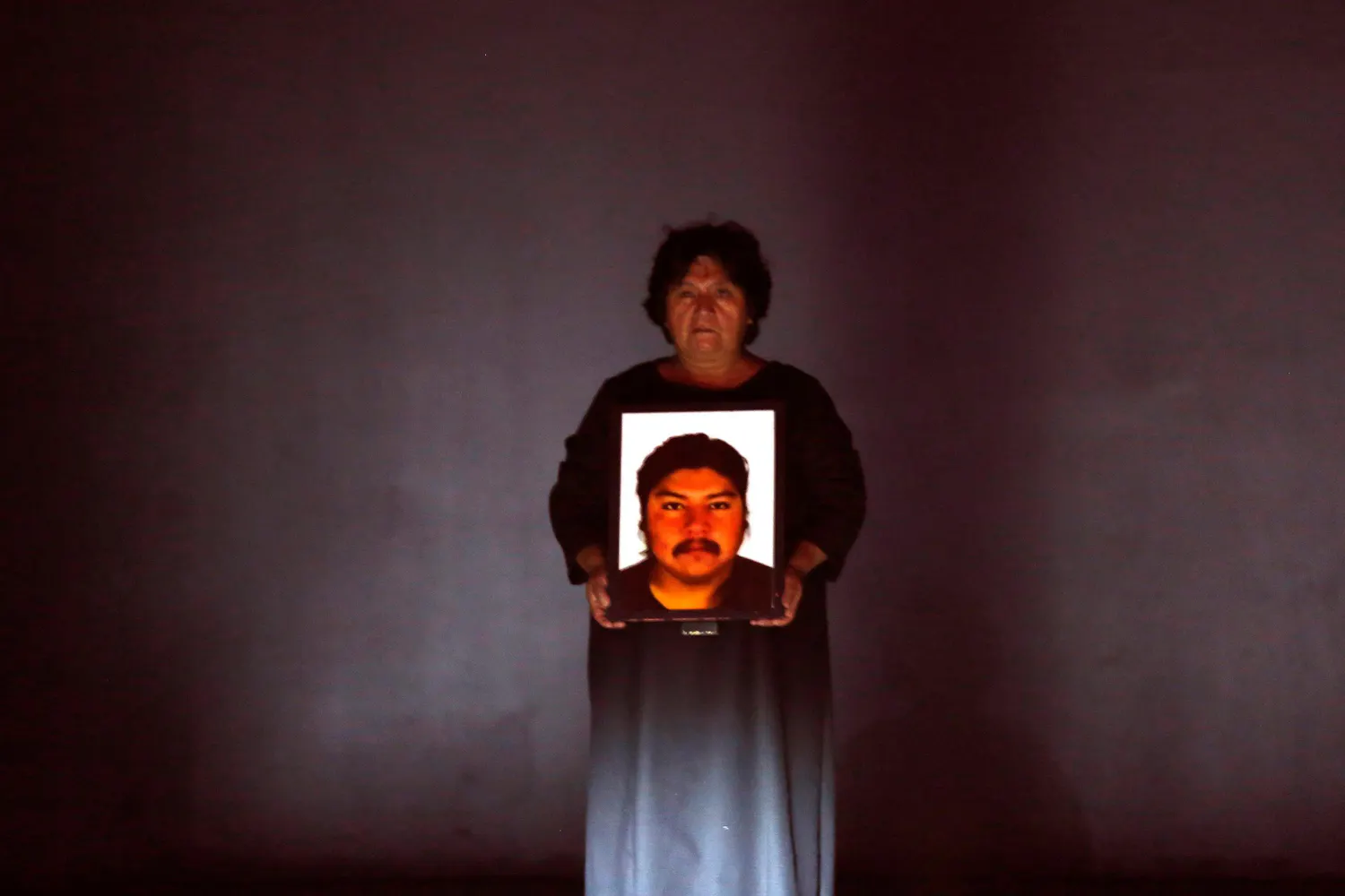 A woman in a gray dress stands on a bare stage staring at the viewer and holding a photo of a man's face.