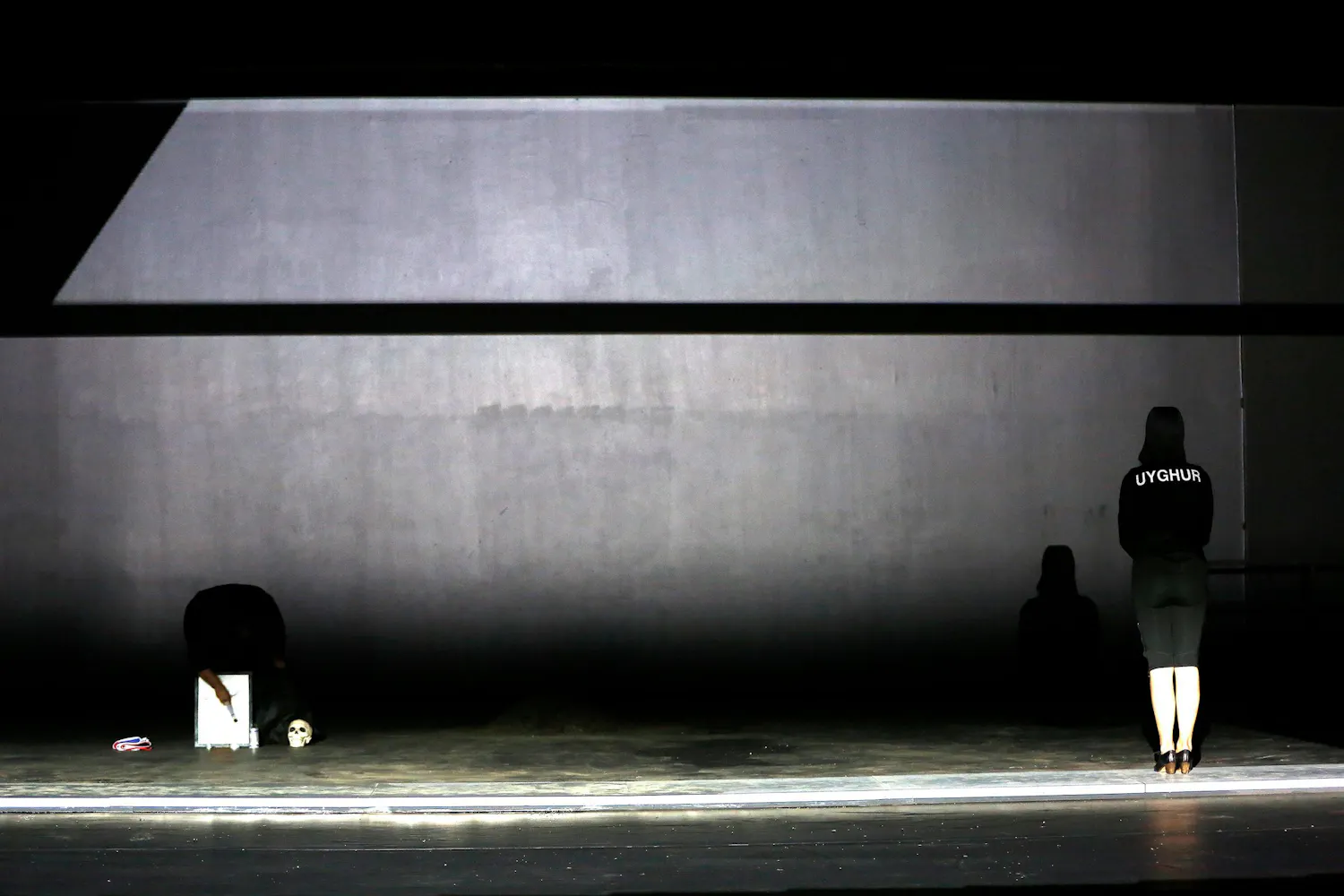 A woman stands on a dark stage with ehr back to the audience in a black hoodie witht he word 