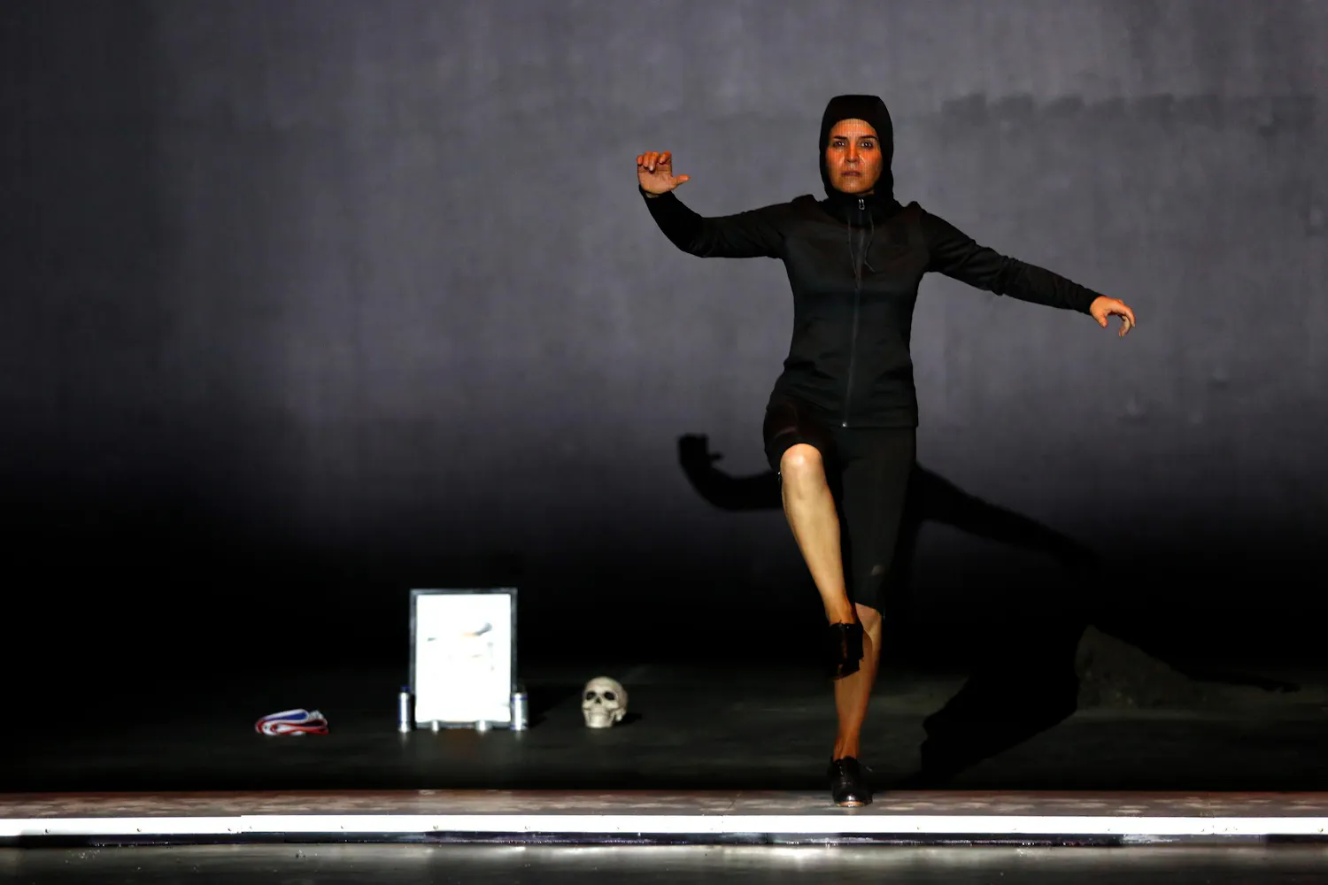 A woman dances on a black stage next to a skull and a framed picture of a man.