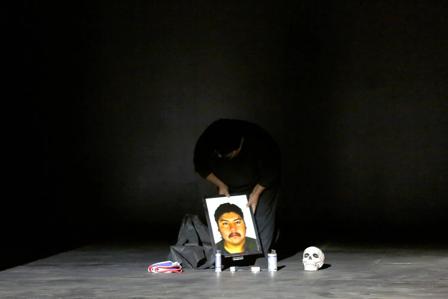 A woman stands on a dark stage setting a framed picture of a man next to candles and a skull.
