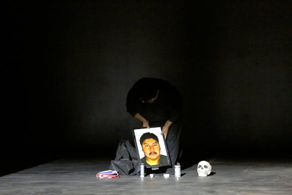 A woman stands on a dark stage setting a framed picture of a man next to candles and a skull.