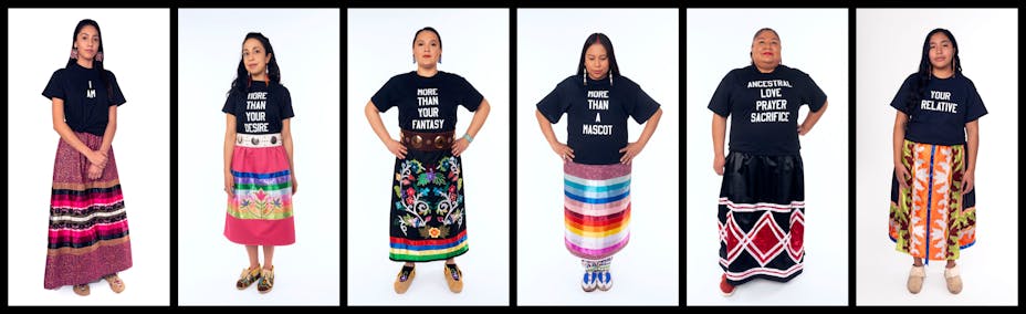 Six indigenous women stand in front of white backgrounds in a horizontal row. each wears a colorful skirt, some wear moccasins, each also wears a black t shirt with white text. In order from left to right they read: I AM, MORE THAN YOUR DESIRE, MORE THAN YOUR FANTASY, MORE THAN A MASCOT, ANCESTRAL LOVE PRAYER SACRIFICE, YOUR RELATIVE