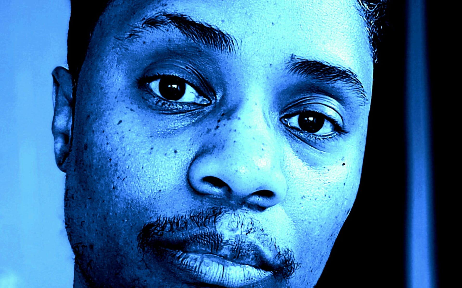 Blue-tinted image of person with short hair, medium skin, and a mustache looking into the camera.