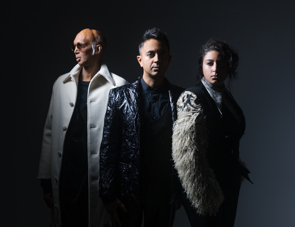 Three adults stand next to one another in a photo studio with one looking away from the viewer and the other two looking at the viewer.