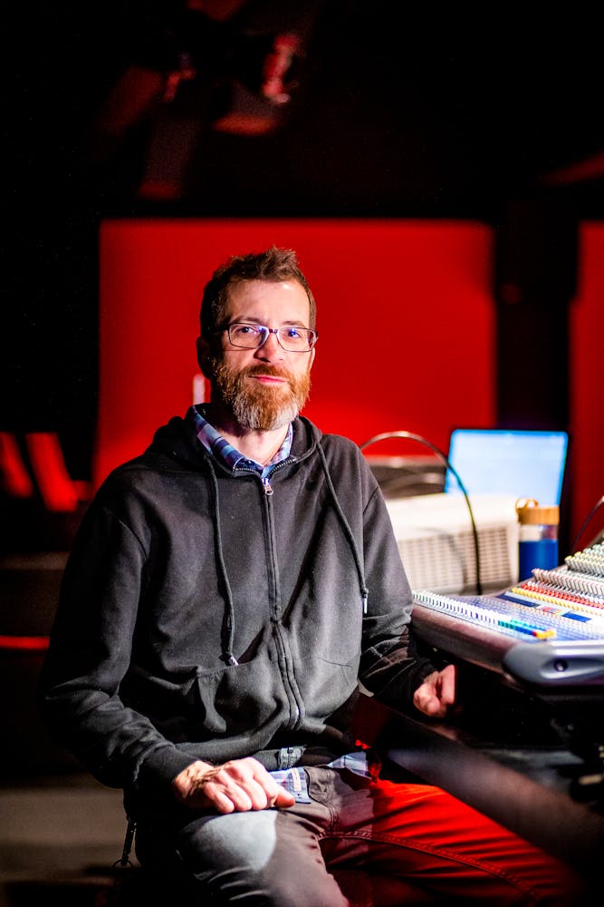 A man sits at a sound mixing board in a cinema and looks at the viewer.