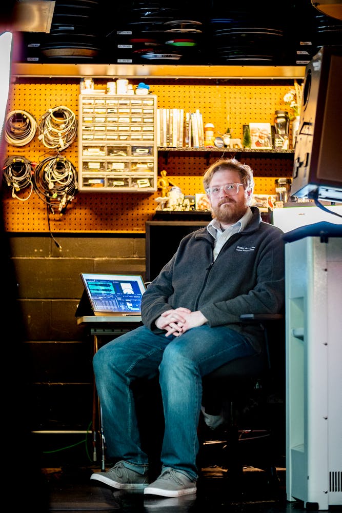 A man sits in a shair in a film projectionist booth and looks at the viewer.