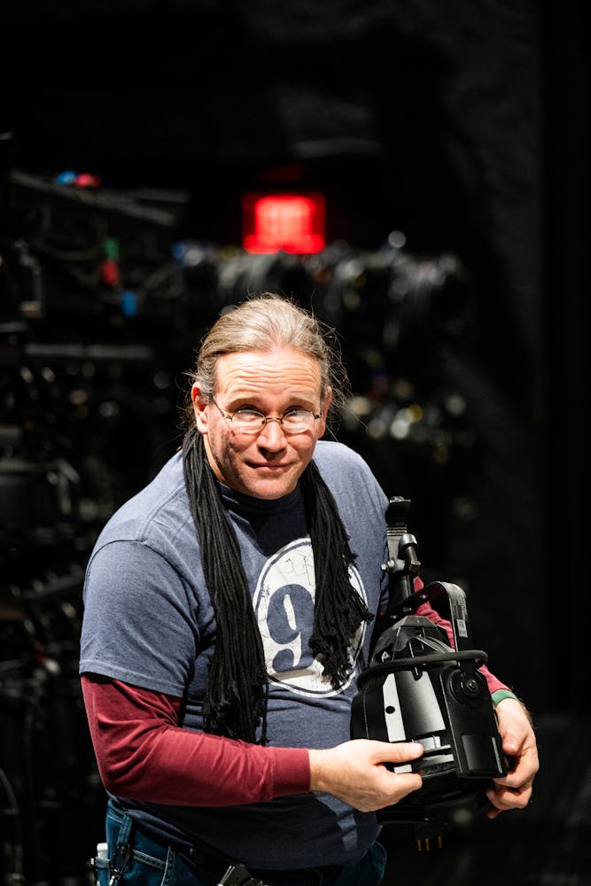 A man with glasses stands on a theater stage holding a light and looks over his glasses at the viewer.
