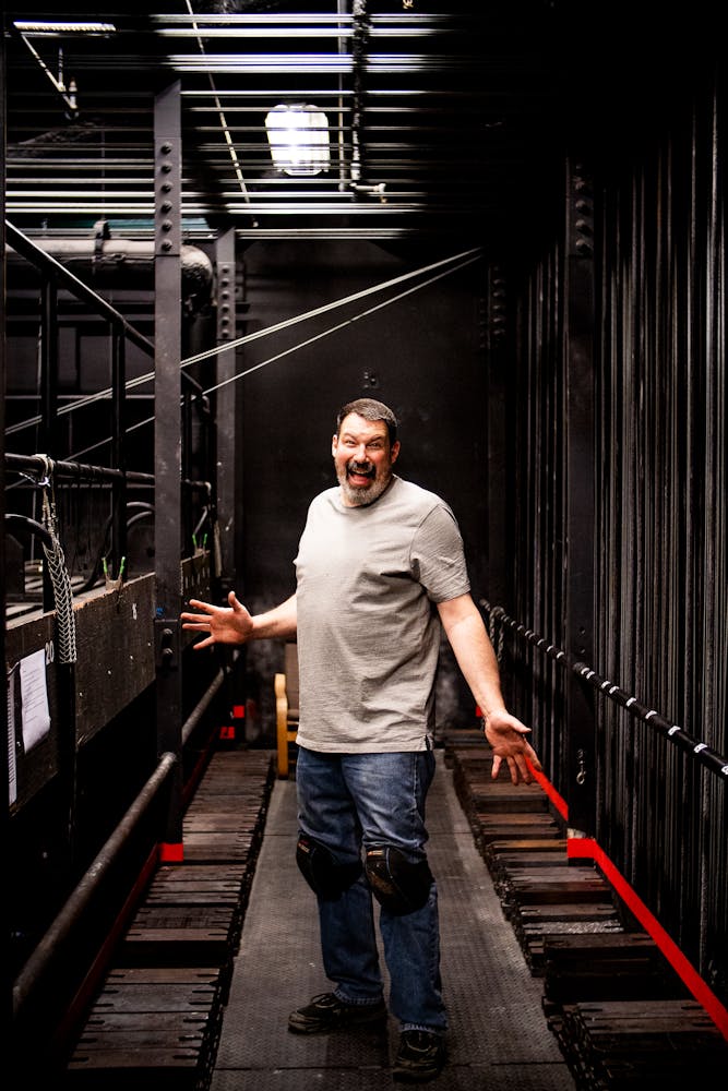A man smiles and holds his arms out at the viewer while standing in the grid over a performing arts theater.