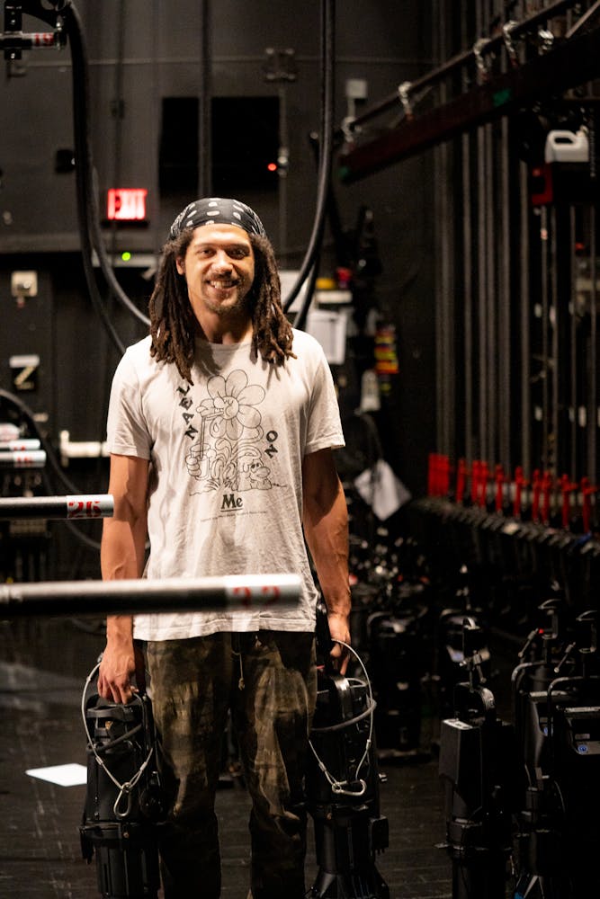 A man stands on a theater stage and smiles at the viewer while holding theaterical lights in each hand.