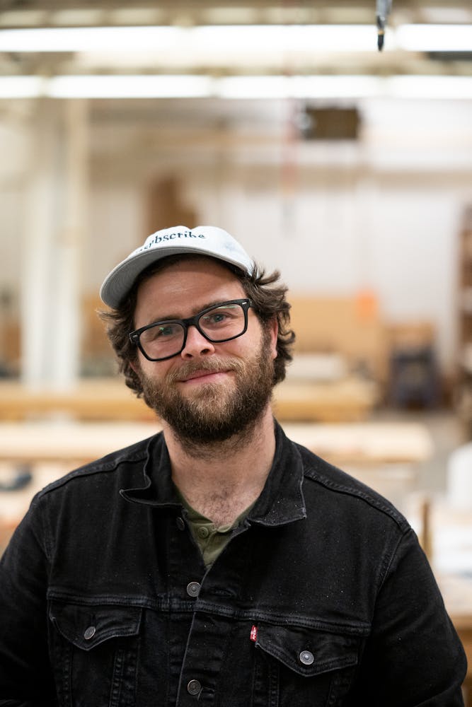 An adult with light skin and a beard and glasses, sits looking at the viewer.