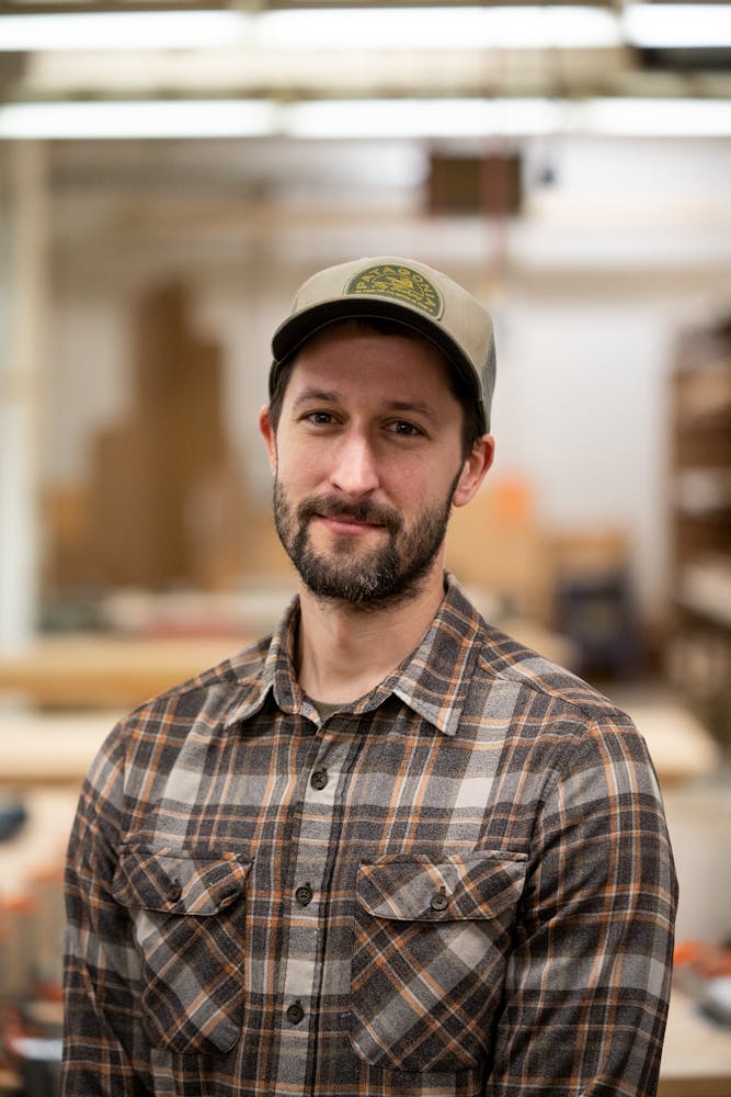 An adult with light skin and dark facial hair, brown flannel shirt and a hat smiles at the viewer.