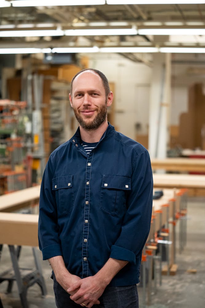An adult with light skin, short hair and a beard stands facing the viewer in a workshop.