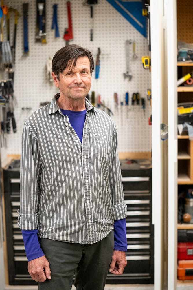 An adult with light skin, brown short hair and a stripped shirt stands facing the viewer in a workshop space.