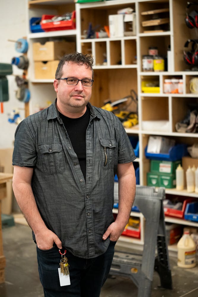 An adult with light skin, brown curly short hair, a grey denim shirt stands facing the viewer in a workshop space.