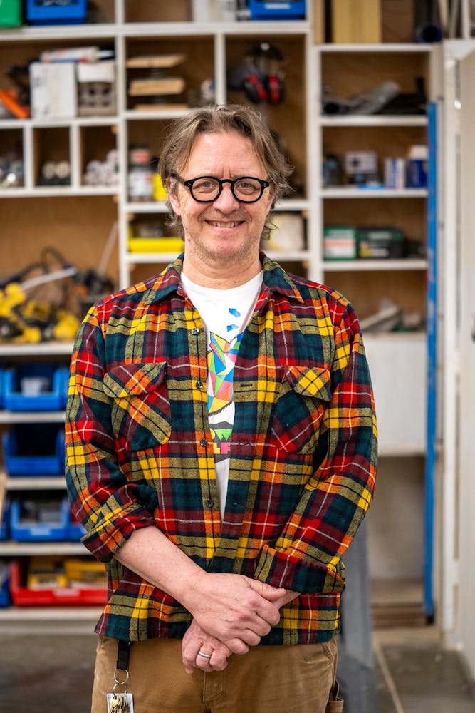 An adult with light skin, black glasses and short blonde hair in a red flannel stands smiling at the viewer in a workshop space.