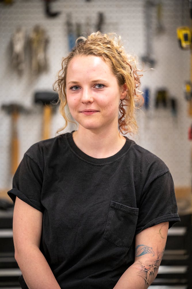 An adult with light skin, black shirt, and long blonde curly hair pulled back faces the viewer, out of focus workshop in the background.