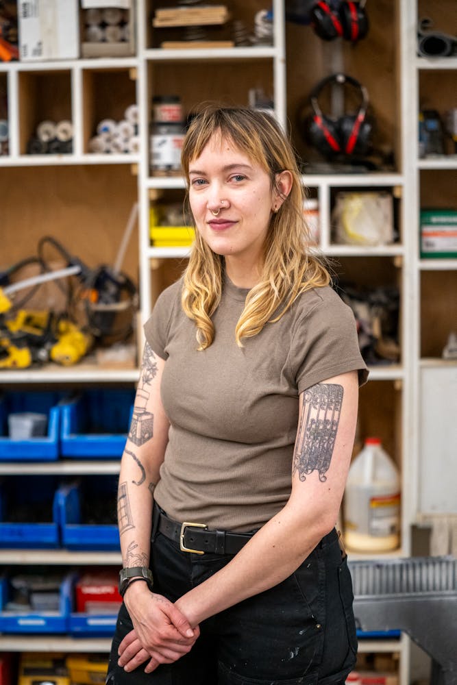 An adult with light skin and long blonde hair with bangs, wearing a brown t-shirt and arm tattoos, stands smiling at the viewer in a workshop space.