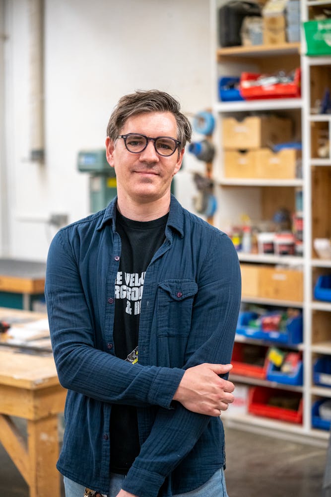 An adult with light skin, a blue long sleave shirt wears dark glasses and looks at the viewer in a workshop space.